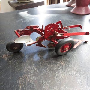 May include: A red toy tractor plow with black rubber tires and a silver blade. The vintage farm equipment model has a detailed design, showcasing the mechanics of a real plow. The toy is a miniature replica of a tractor plow.