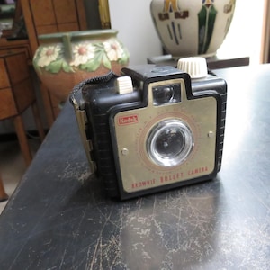 May include: A vintage Kodak Brownie Bullet camera. The camera is black with a gold-colored faceplate and a white dial. The Kodak logo is visible. The camera has a strap and is sitting on a dark surface.