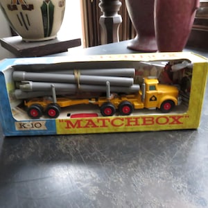 May include: A vintage Matchbox K-10 truck toy in its original box. The yellow truck carries gray pipes on a flatbed trailer. The box is blue and yellow with the word "MATCHBOX" in red.