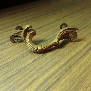 May include: A gold-toned metal drawer pull with a decorative, ornate design. The pull has a curved shape with a scroll-like pattern.