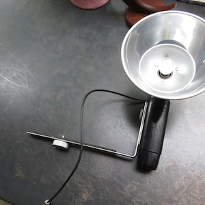 May include: A vintage photographic flash unit with a silver reflector and a black handle. The flash head is round and has a bulb in the center. A black cord extends from the unit. The flash is mounted on a metal bracket.