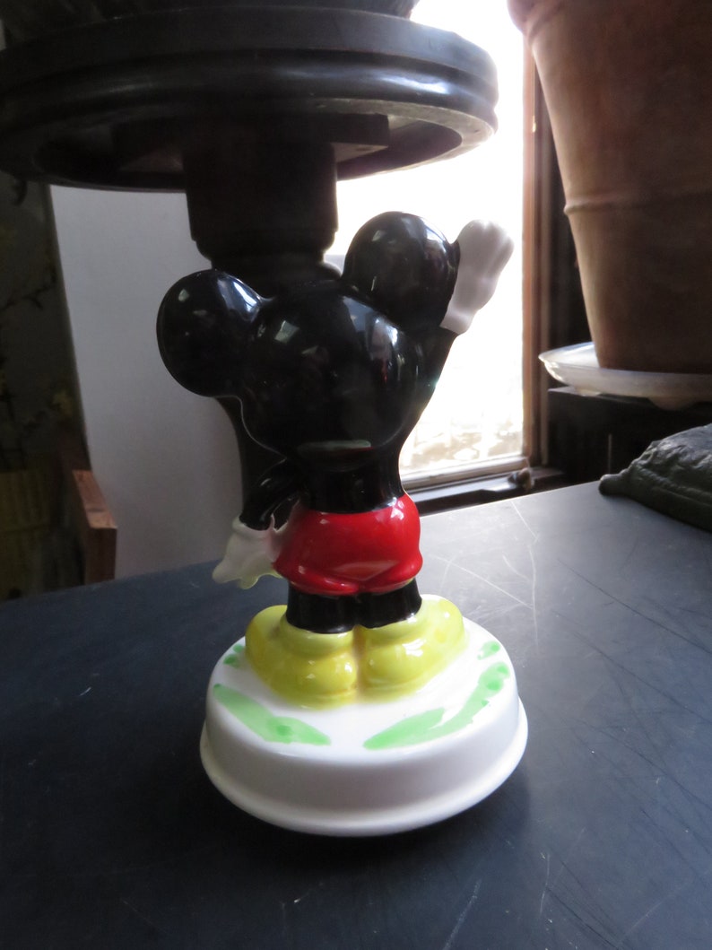 May include: A vintage wind-up music box featuring a black and white Mickey Mouse figurine with red shorts and yellow shoes. The music box is white with green accents and is on a black surface.