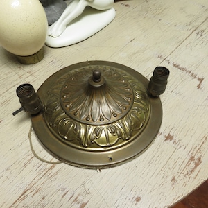 Op de afbeelding: Een vintage messing plafondlamp met twee fittingen. De armatuur heeft een sierlijk, bloemmotief op de centrale plaat. De lamp is rond en heeft een centrale decoratieve finial.