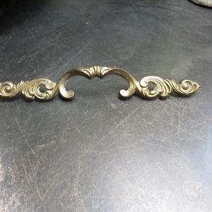 May include: Ornate brass-toned cabinet pull with a curved handle and decorative scrollwork on each end. The hardware is designed for furniture and features a classic, vintage aesthetic. The pull is approximately 6 inches long.