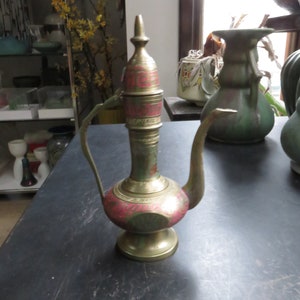 May include: A gold and red metal teapot with a decorative design. The teapot has a round body and a long spout. The teapot is sitting on a black surface.