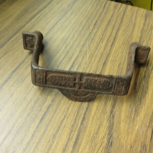 May include: A rusty metal U-shaped bracket with an ornate design. The bracket has a decorative pattern of leaves and swirls.