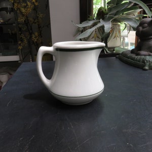 May include: White ceramic creamer with a curved body and a handle. A thin green line accents the rim and base. The creamer is on a dark surface. The background includes a window and decorative items.
