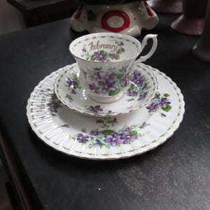 May include: A vintage teacup, saucer, and plate set. The white porcelain features a purple floral design with gold trim. The teacup has the word "February" printed on it.