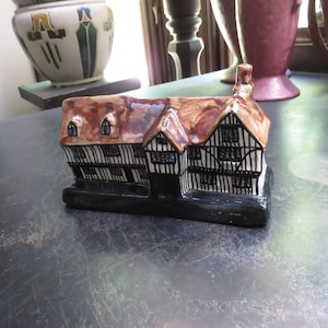 May include: A ceramic miniature cottage figurine featuring a brown glazed roof, black and white timber-framed walls, and a solid black base. The structure includes multiple small windows and a chimney, designed with a glossy finish throughout.