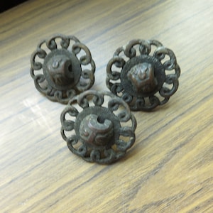 May include: Three antique bronze cabinet knobs with ornate floral designs and a raised center detail. The knobs are on a wooden surface.