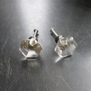 May include: Two clear glass cabinet knobs with a hexagonal shape and a silver screw. The knobs have a gold-colored metal insert in the center.