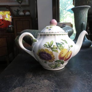 May include: White ceramic teapot with a floral design featuring sunflowers and yellow fruit. The teapot has a pink knob on the lid and a curved handle. The design includes green leaves and a scalloped edge.