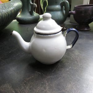 May include: A small, white enamel teapot with a lid and a dark blue handle. The teapot has a curved spout and a black rim. Other ceramic items are in the background.