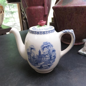 May include: White ceramic teapot with a blue and white landscape design. The teapot has a curved spout and handle, and a lid topped with a pink rose. The design features a building and trees.