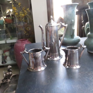 May include: A silver tea set with a teapot, creamer, and sugar bowl. The set is on a dark wooden table.