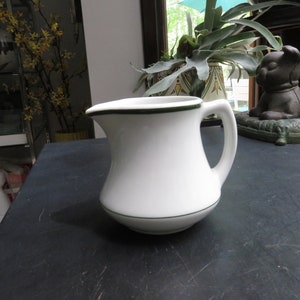 May include: White ceramic creamer with a green rim. The creamer has a rounded body and a handle.