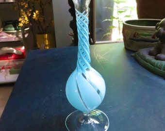 May include: A light blue glass vase with a twisted design and a clear glass base. The vase is sitting on a dark blue surface.