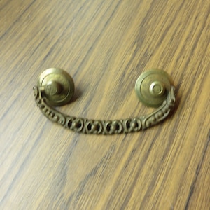 May include: A brass drawer pull with a decorative, ornate design. The pull has two round knobs on either end, connected by a curved, detailed bar.