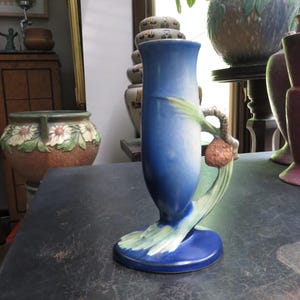 May include: A blue ceramic vase with a flared top and a wide base. The vase features a green and brown decorative element on the side. The vase is displayed on a dark surface.