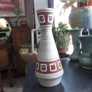 May include: A white ceramic vase with a brown and orange geometric pattern. The vase has a handle and a wide base.