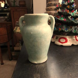 May include: A light blue ceramic vase with two handles. The vase has a slightly textured surface and a rounded shape. The vase is displayed on a dark surface, with a ceramic Christmas tree and other decorative items in the background.