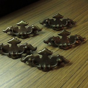 May include: Five antique brass drawer pulls with ornate designs. Each pull has a decorative, leaf-shaped design and a simple, round handle.