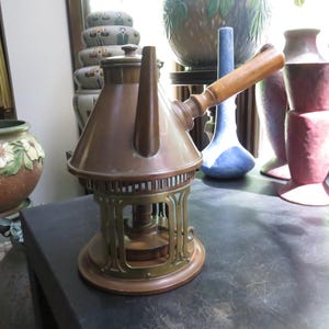 May include: A copper-colored metal tea kettle with a conical body, a long wooden handle extending from the side, and a brass-toned openwork base featuring vertical slats. The kettle has a flat lid and a central spout, resting on a flat surface.