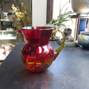 May include: A red glass pitcher with a yellow amber base and a clear glass handle. The pitcher has a rounded body and a wide mouth.