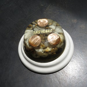 May include: A clear dome paperweight with three copper-colored coins embedded in a bed of white and black stones. The words "LAS VEGAS" are written in gold lettering on the dome. The paperweight is sitting on a white base.