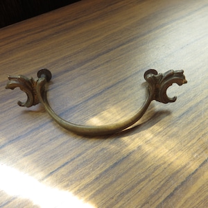 May include: A brass drawer pull with ornate scrollwork on the ends. The pull is curved and has a smooth, polished finish.