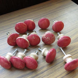 May include: A collection of 15 red and white wooden drawer knobs. The knobs have a distressed finish and are arranged on a wooden surface.