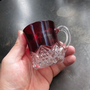 May include: A small, ruby-red glass mug with a clear handle and a diamond-cut base. The mug has text etched on the side. The mug is held in a person's hand. The mug is likely vintage or antique.