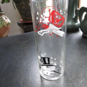 May include: A clear glass drinking glass with a white and red design. The design features a top hat, a pair of binoculars, a necklace, and a red and white patterned object.
