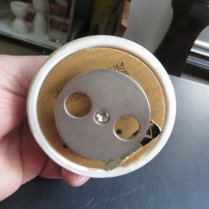 May include: A white ceramic music box with a wooden base. The music box has a metal winding mechanism on the bottom. The base has the text "Made in Japan" printed on it.