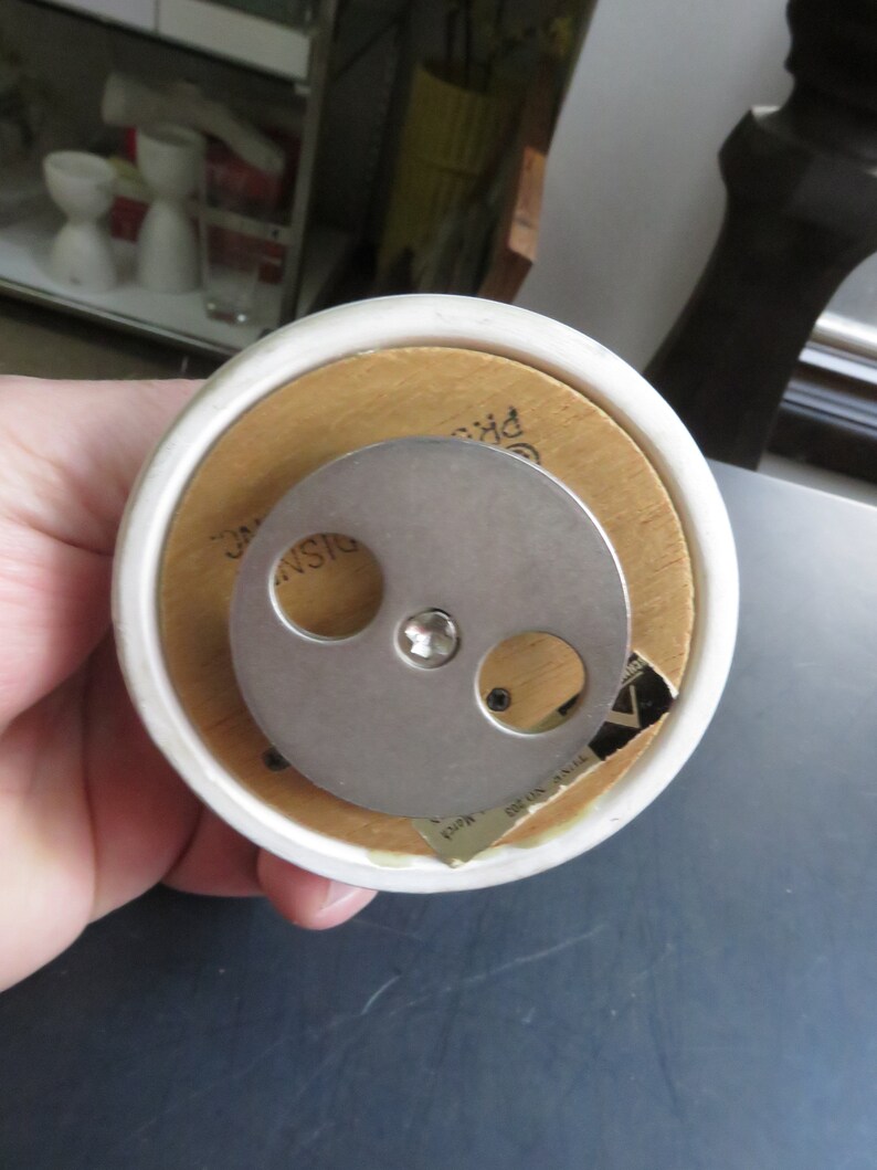 May include: A white ceramic music box with a wooden base. The music box has a metal winding mechanism on the bottom. The base has the text "Made in Japan" printed on it.