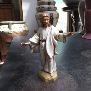 May include: A ceramic figurine of Jesus Christ, depicted with long brown hair and a beard, wearing a white robe and a draped shawl. The figure stands on a small, tan-colored base with arms outstretched. The statue is a religious item.