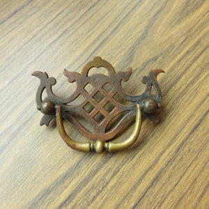May include: A vintage brass cabinet pull with a decorative, openwork design. The pull has a D-shaped handle and two round knobs.