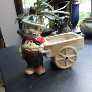 May include: A ceramic figurine of a man wearing a green hat and red scarf, pushing a small, beige wheelbarrow planter. The planter has a white interior and a brown rim.
