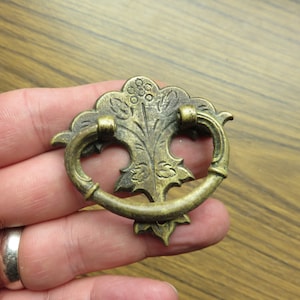 以下が含まれることがあります： 装飾的な花のデザインが施された真鍮製のキャビネットプル。プルはリング型で、家具に使用するために設計されています。