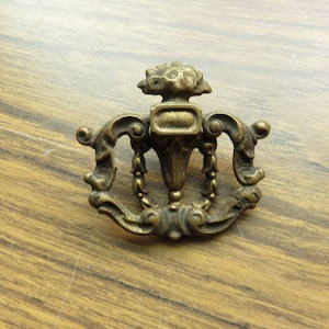以下が含まれることがあります： 中央に壺、スクロール模様が施された、装飾的なデザインのヴィンテージ真鍮製の引き出しノブです。