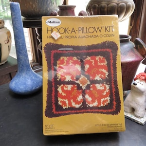 May include: A rectangular Malina Hook-A-Pillow kit box with a yellow background. The box features a graphic of a square pillow with a dark brown border and a central floral pattern in orange, red, and white. Text includes Hook-A-Pillow Kit, 12"x12", and Style #30/23 Spanish Tile.