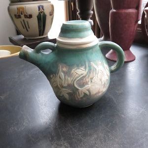 May include: A teal ceramic teapot with a lid and handle. The teapot features a white band around the lid and a floral design on the body. The spout is on the left side of the teapot.