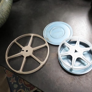 May include: Three vintage film reels and a light blue film canister are displayed on a dark surface. The reels are silver-colored, with a starburst design. The canister has a matching light blue lid.