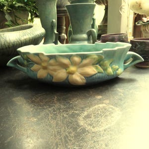 May include: A turquoise ceramic planter with a floral design. The planter has two handles and is decorated with yellow and white flowers. Other ceramic vases and planters are in the background.