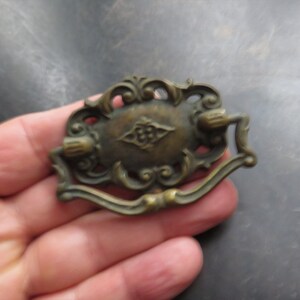 May include: A vintage brass cabinet pull with an ornate design. The pull has a diamond-shaped center with a floral pattern and two decorative scrolls on either side.