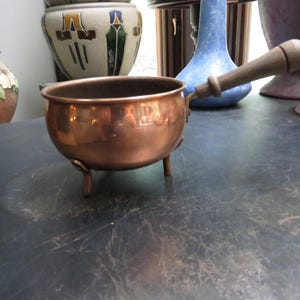 May include: A small, round copper pot with a polished metallic finish, featuring three short legs for stability and a single, long, cylindrical wooden handle attached to the side. The interior of the pot is dark, and the exterior surface is smooth.