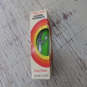 May include: A vintage box of Looney Tunes Anti-Cavity Fluoride Toothpaste with a cartoon image of Bugs Bunny. The box is green, yellow, orange, and red with the text "Fruity Flavor" and "NET WT. 2.7 OZ."