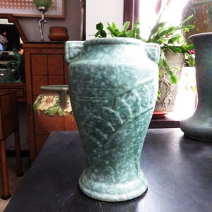 May include: A green ceramic vase with a speckled glaze and a raised floral design. The vase has a rounded body and a flared lip.