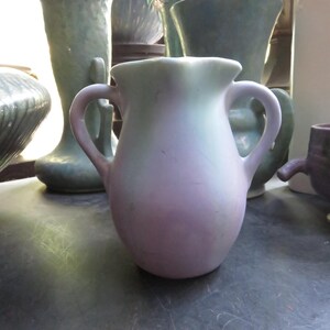 May include: A small, two-handled vase with a rounded body and a ruffled rim. The vase has a gradient color scheme, transitioning from a soft lavender at the base to a pale green at the top. Other vases are visible in the background.