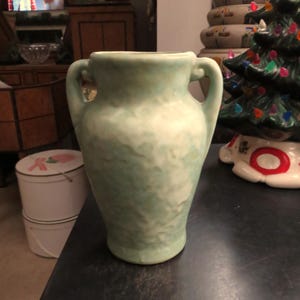 May include: A light green ceramic vase with two handles. The vase has a textured surface and a slightly flared rim. The vase is sitting on a dark surface.